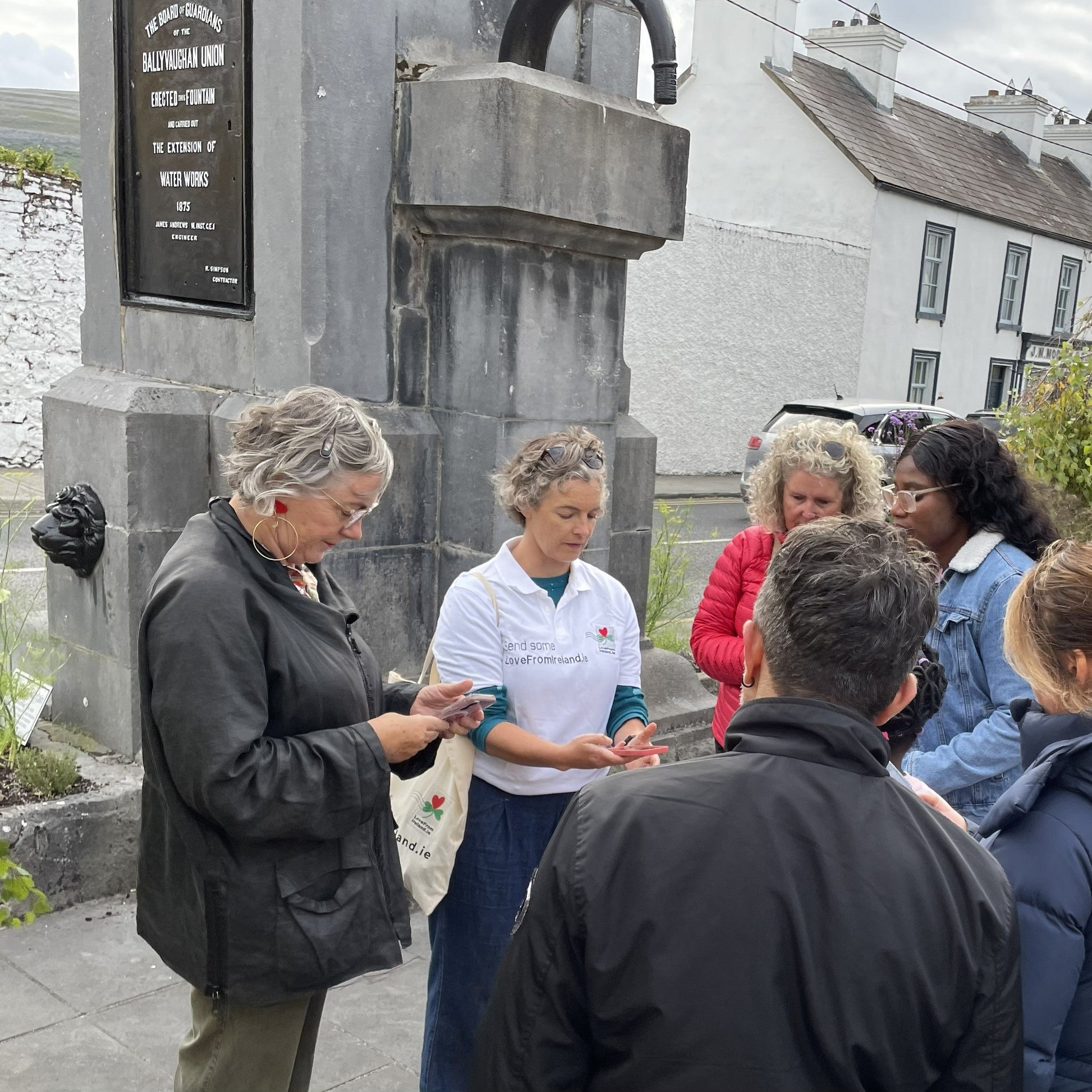 A group of photowalk participants standing beside the fountain in Ballyvaughan village as Claire O'Rorke guides them through the advanced camera settings on their phones.