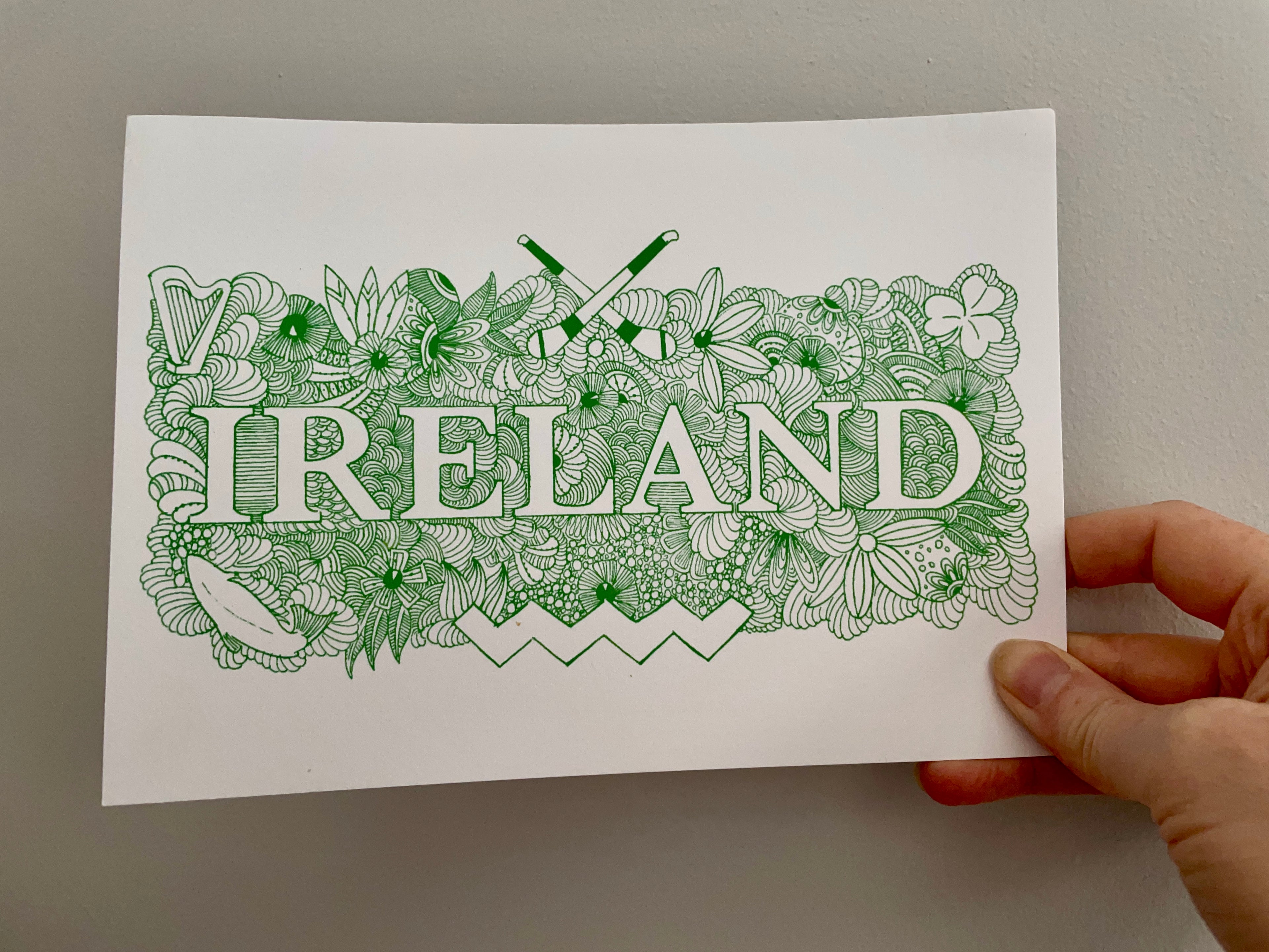 A hand holding the beautifully embellished "Ireland" postcard by Audrey Cantillion featuring the word Ireland illuminated with lots of Irish cultural items such as hurleys, a harp, a shamrock, flowers and seashells. It is in green ink.